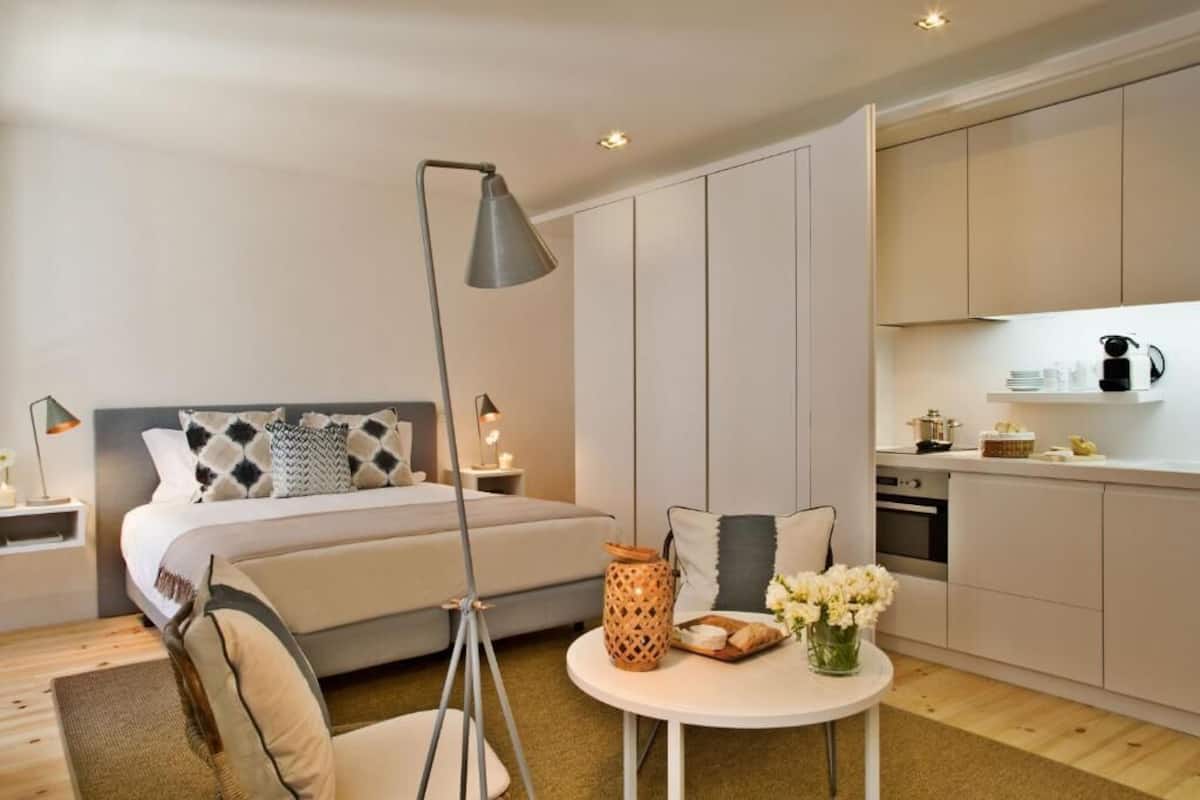 Een moderne hotelkamer met een bed, nachtkastje, lamp en een kleine eettafel met bloemen.