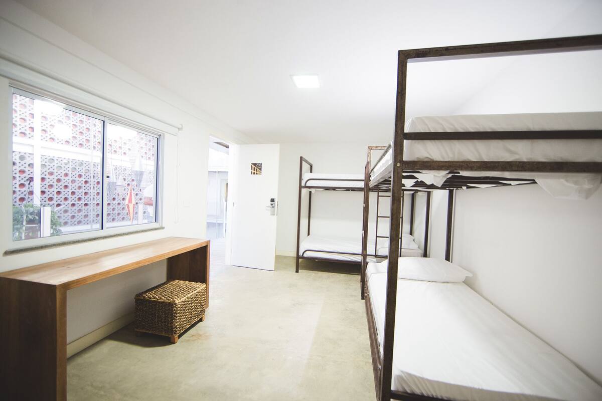 A dormitory room with bunk beds, a window with a view, a wooden bench, and a small woven basket.