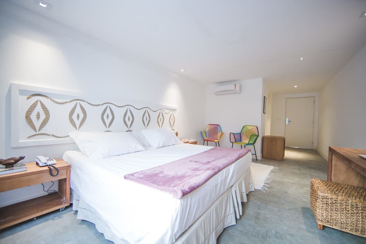 A bedroom with a large bed, a bedside table with a phone, a chair, a small wooden cabinet, and a wall-mounted air conditioner.