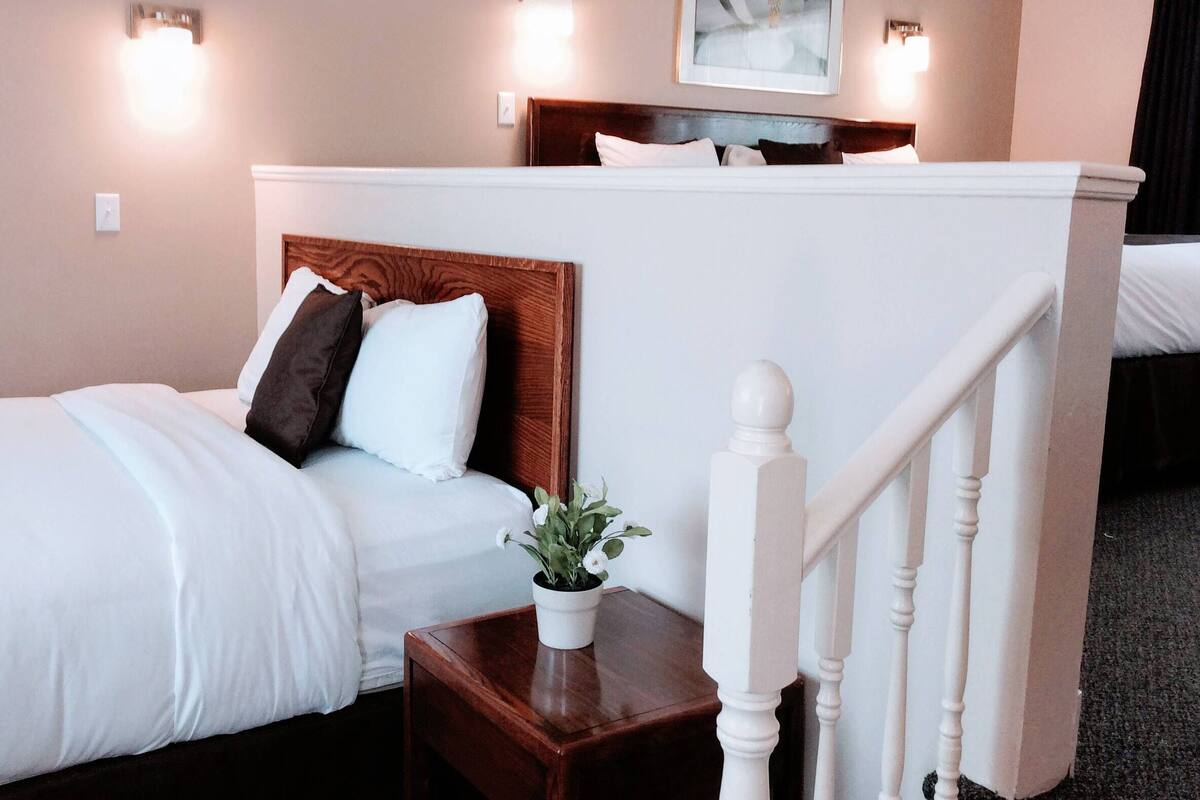 A hotel room with a wooden headboard bed, a small bedside table with a plant, and a white railing.