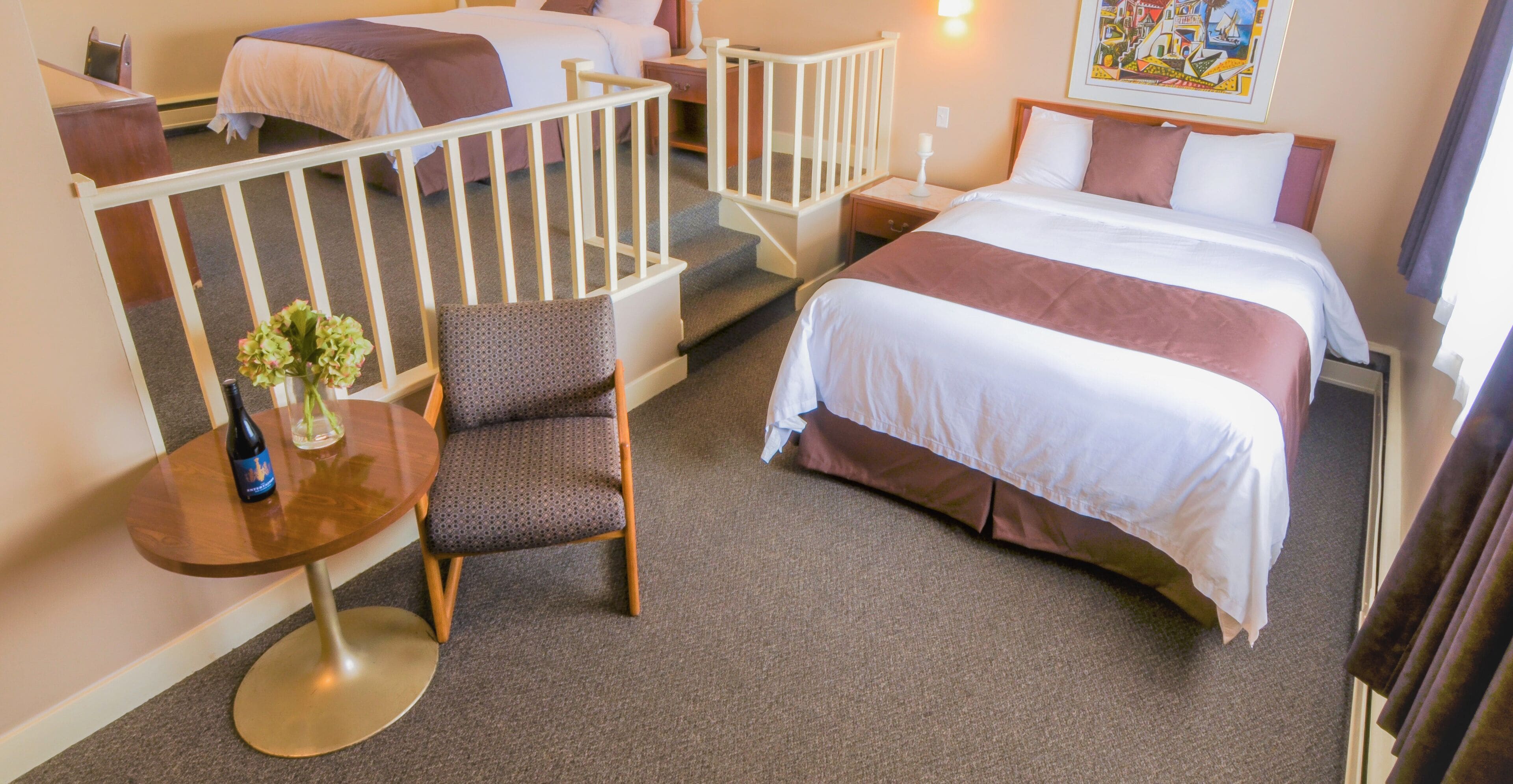 A hotel room with a large bed, a smaller bed, a chair, a table with a vase, and a staircase.