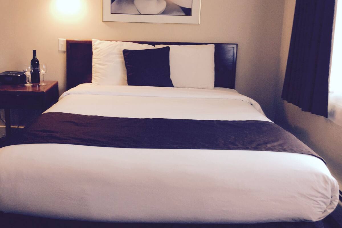 A neatly made bed with white and dark bedding, a small bedside table with a bottle and glass, and a framed picture on the wall.