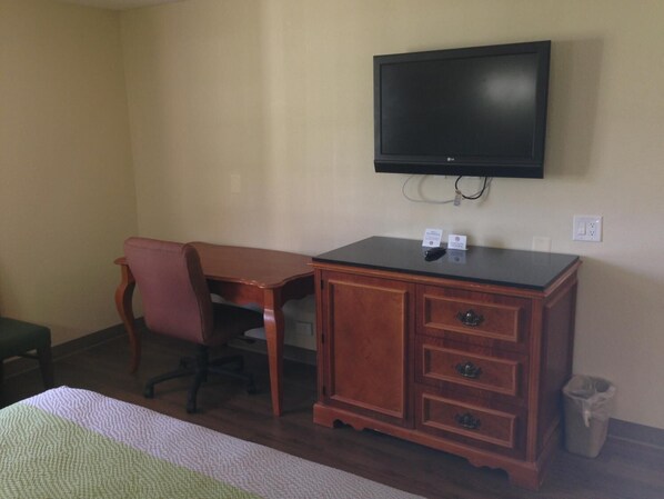 A hotel room with a wooden desk, a chair, a television mounted on the wall, and a trash can.