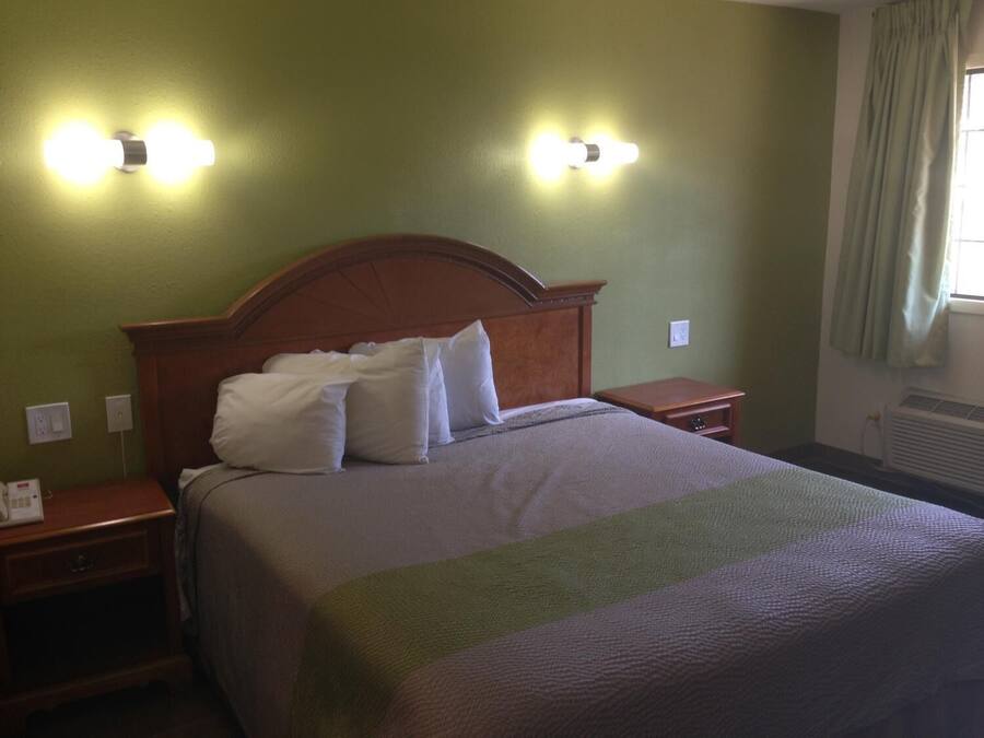 A hotel room with a wooden headboard, a bed with white pillows, two bedside tables, and wall-mounted lights.