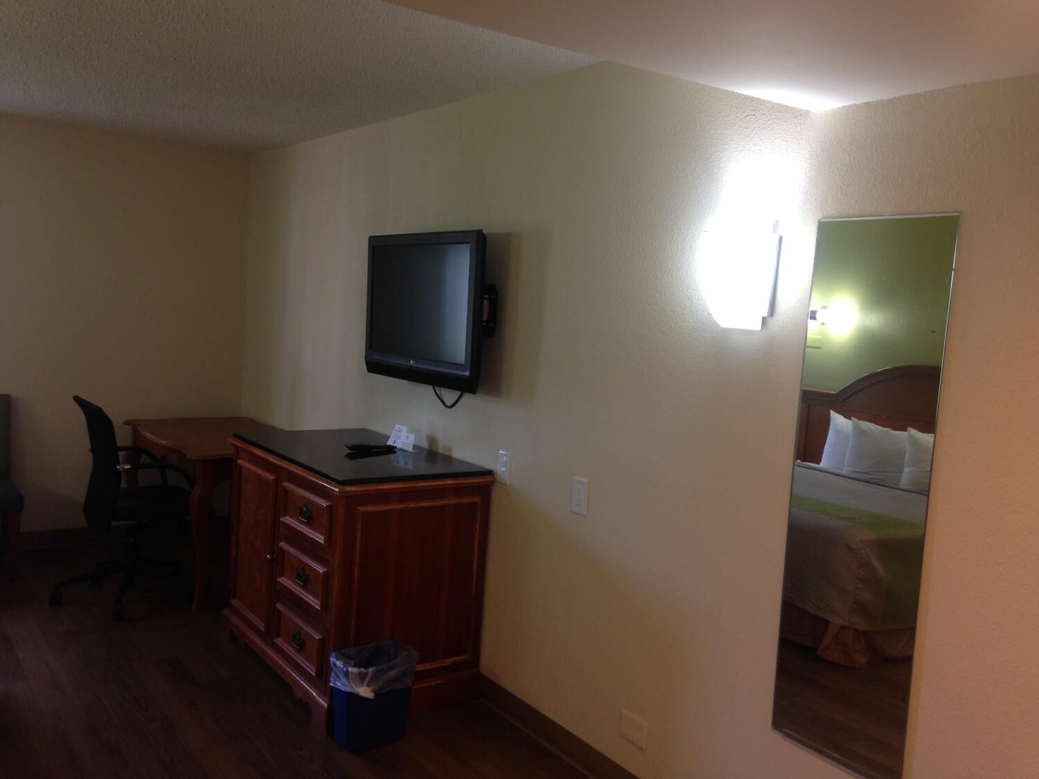 Una habitación de hotel con un escritorio de madera, un televisor montado en la pared, un cubo de basura y una cama con pared verde y ropa de cama blanca.