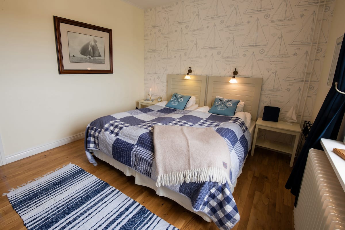 A bedroom with a bed, a nightstand, a bedside table, a framed picture on the wall, and a patterned wallpaper.