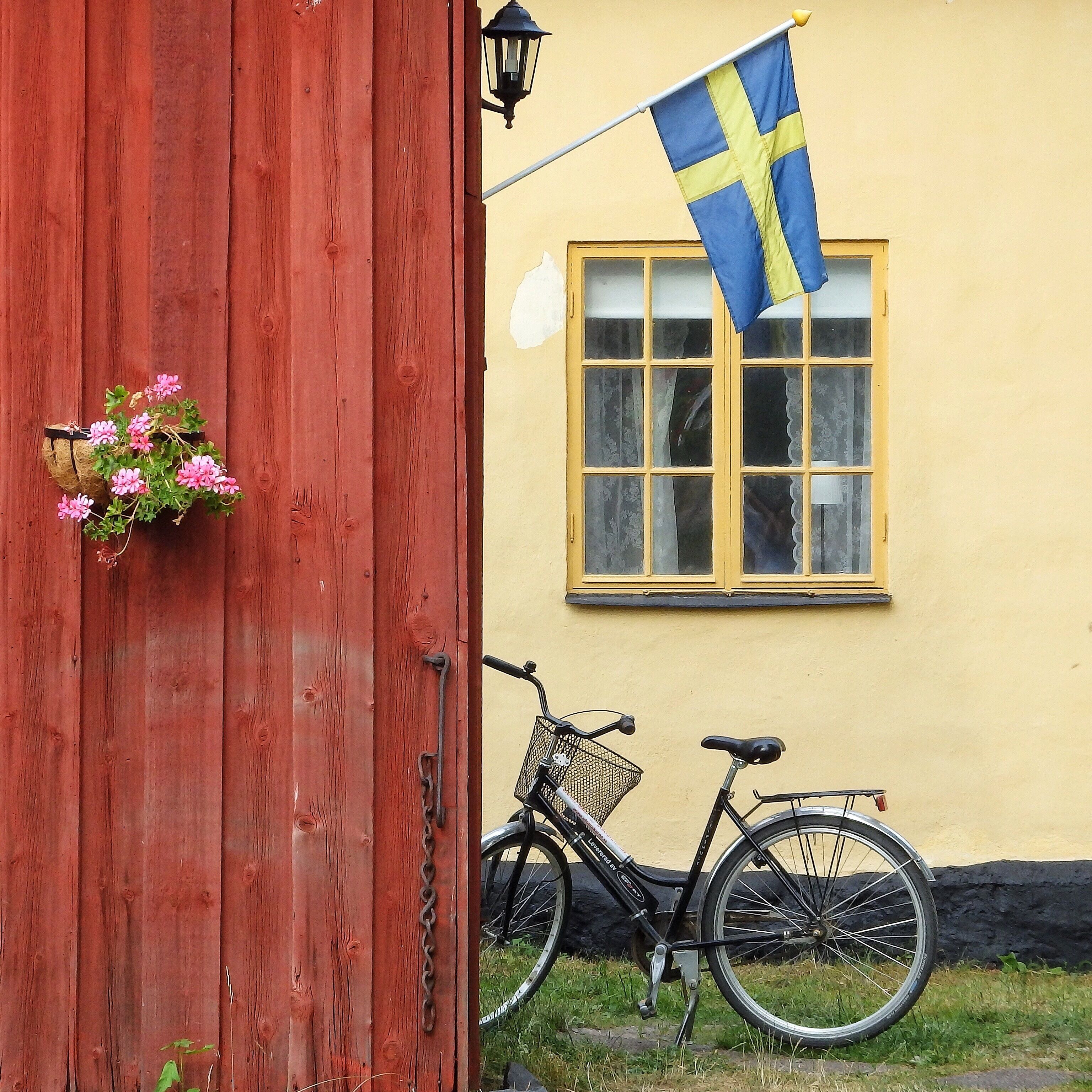 En röd trädörr med en blomkorg, en svensk flagga och en cykel parkerad utanför en gul byggnad med ett fönster.