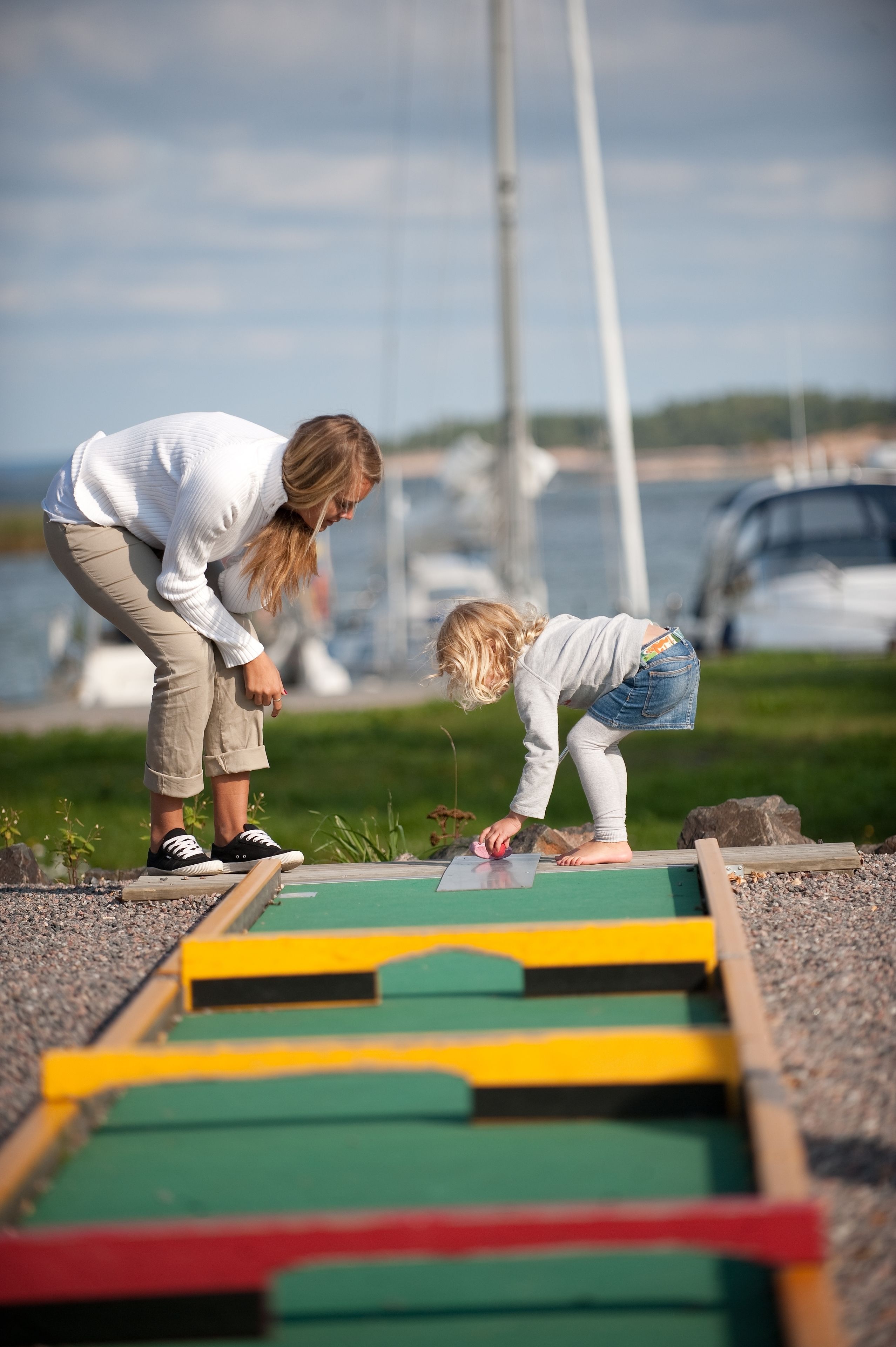 Deux personnes jouant à un jeu de société à l’extérieur, avec un port de plaisance et des bateaux en arrière-plan.