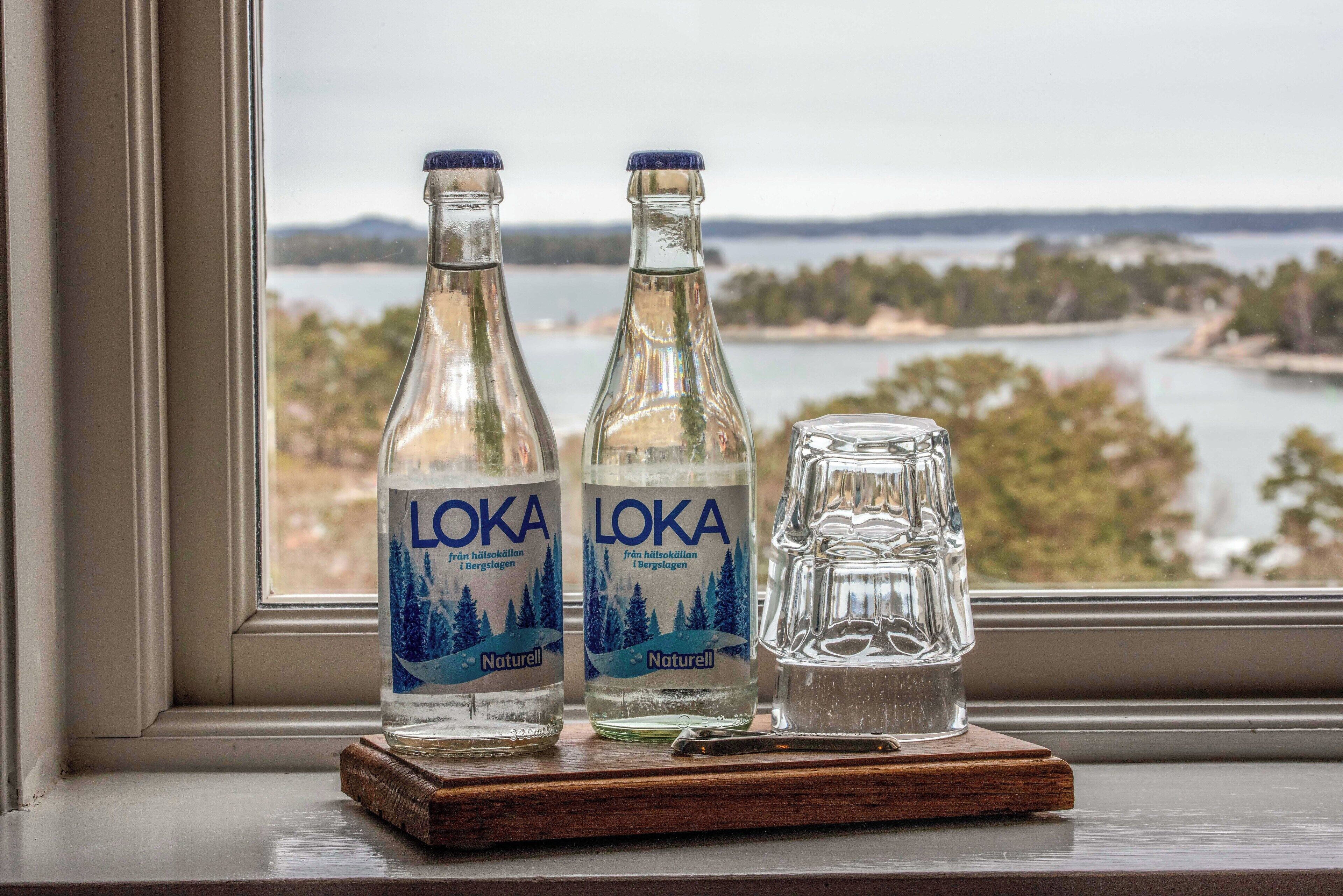Dos botellas de agua LOKA Naturell y un vaso sobre un posavasos de madera, junto a una ventana con vistas panorámicas.