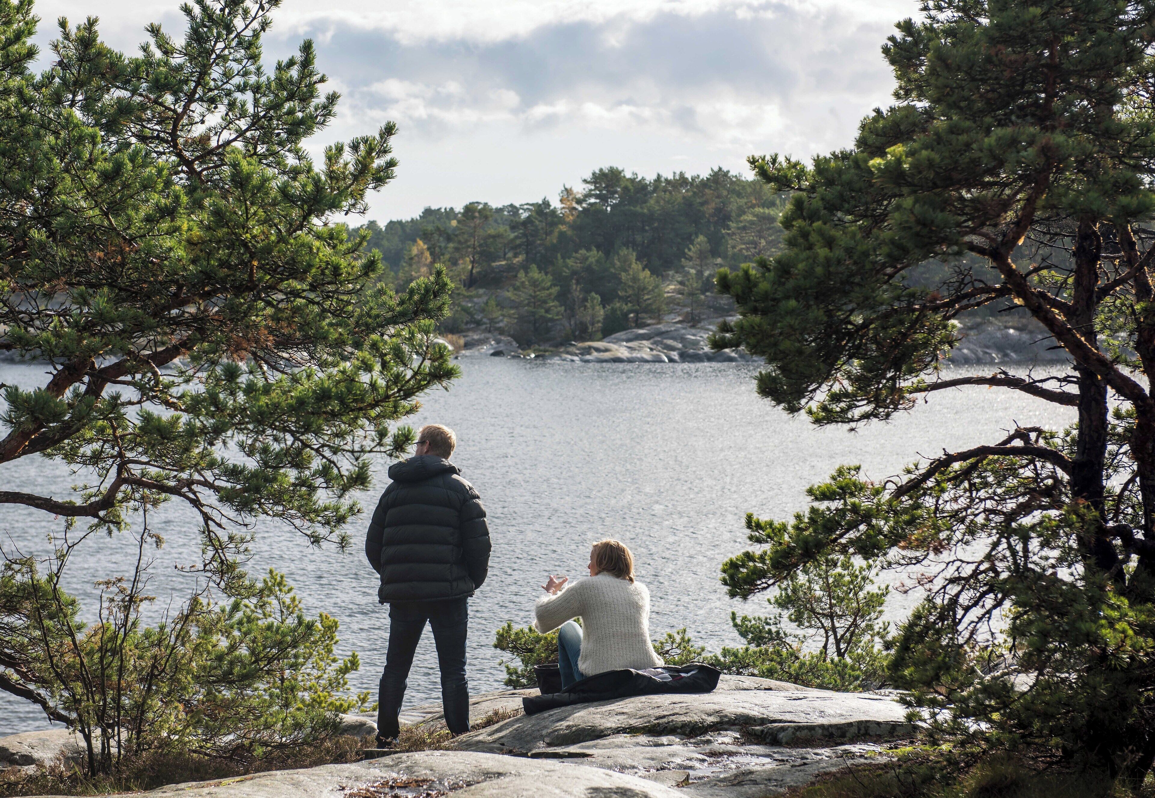Two people sitting on a rock by a lake, surrounded by pine trees.
