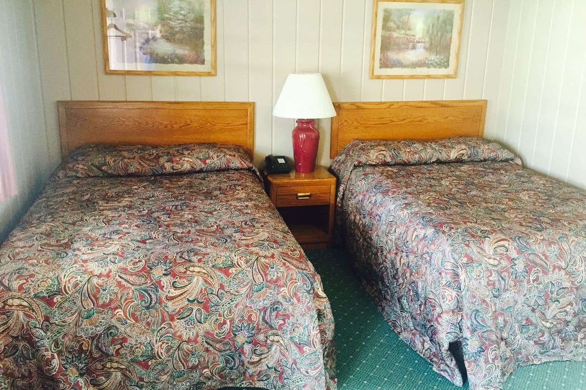 A double room with two beds, each with a patterned bedspread, a bedside table with a lamp, and two framed pictures on the wall.