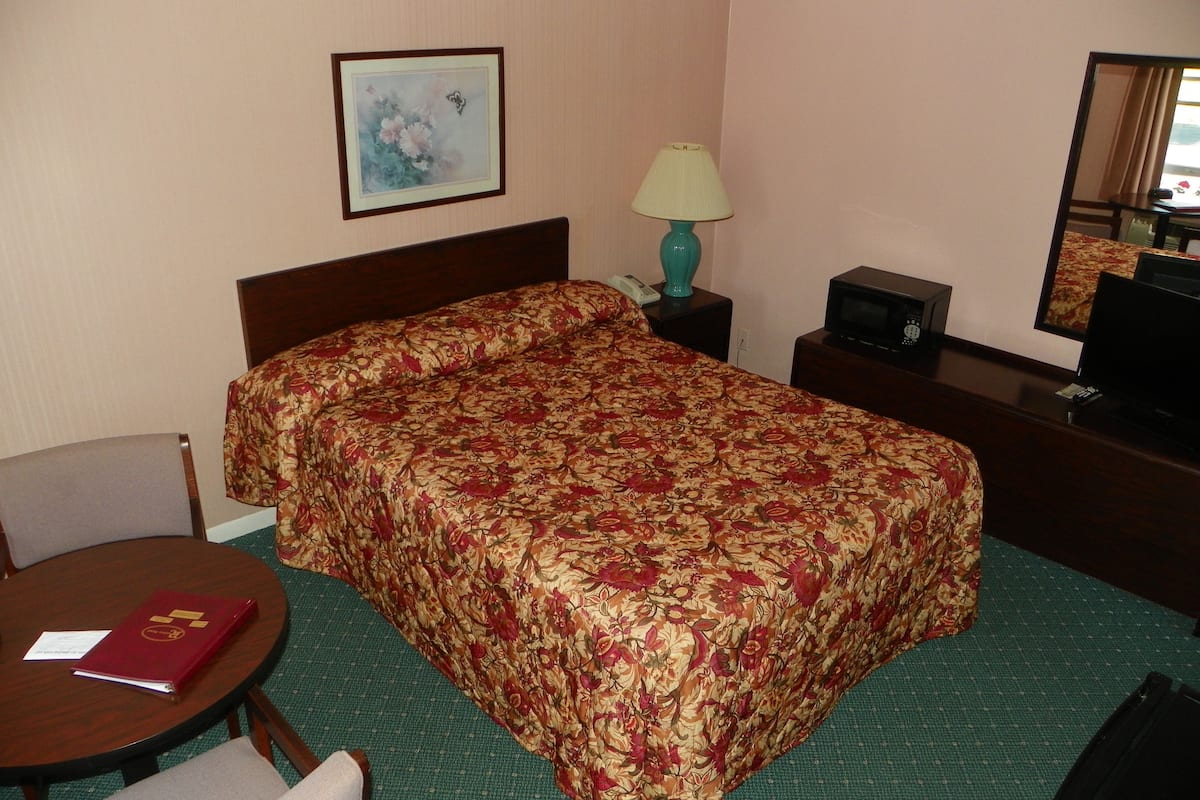 A hotel room with a bed, a round table with a red folder, a lamp, a small table with a remote control, and a mirror.
