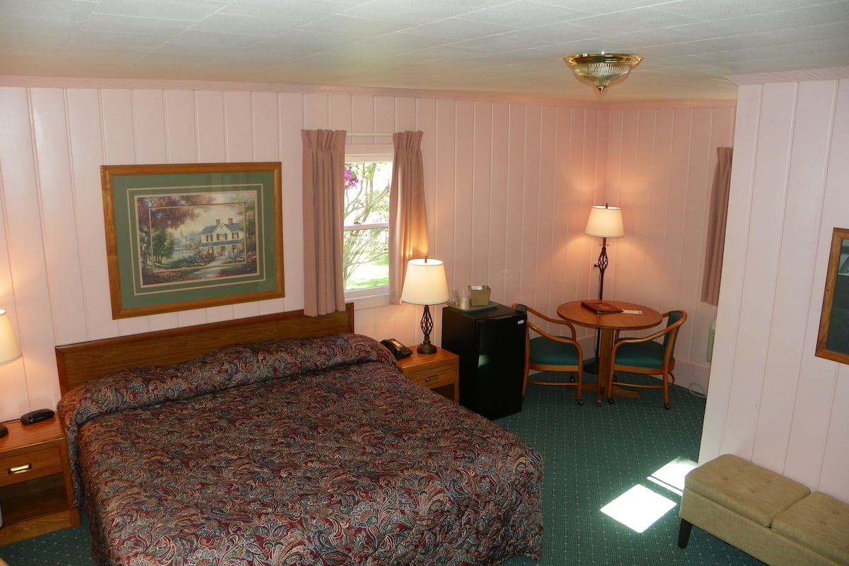 A hotel room with a large bed, a desk with a lamp, a small table with chairs, and a painting on the wall.