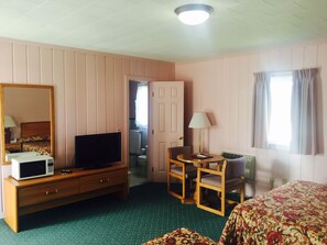 Une chambre d’hôtel comprenant un lit, un bureau avec une chaise, une télévision, un miroir et une fenêtre avec des rideaux.