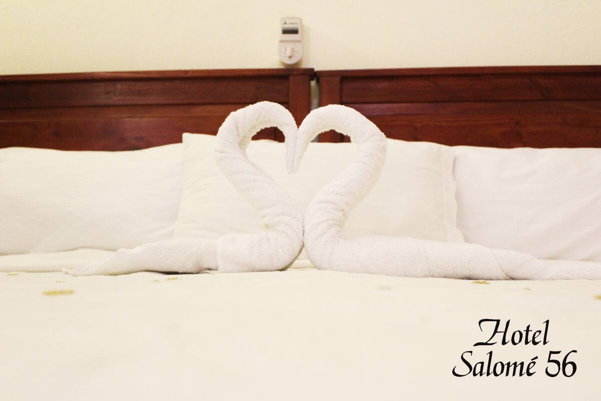Een bed met witte lakens en twee handdoeken die in een hartvorm zijn gerangschikt. De tekst 'Hotel Salomé 56' is zichtbaar.