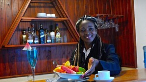 Iemand zit aan een houten bar met een bord fruit, een cocktailglas en een beker op de toonbank. Achter de persoon staat een plank met flessen en glazen.