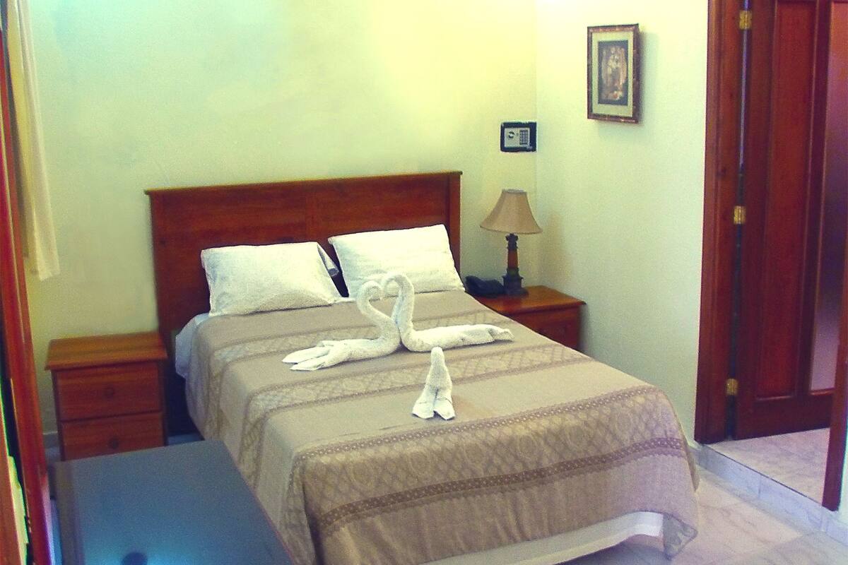 Een netjes opgemaakt bed met een zwaanvormige handdoekdecoratie, een bedlampje en een klein klokje op het nachtkastje.