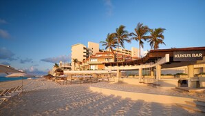 A beachfront hotel with a bar named 'Kukus Bar', palm trees, and a sandy beach.