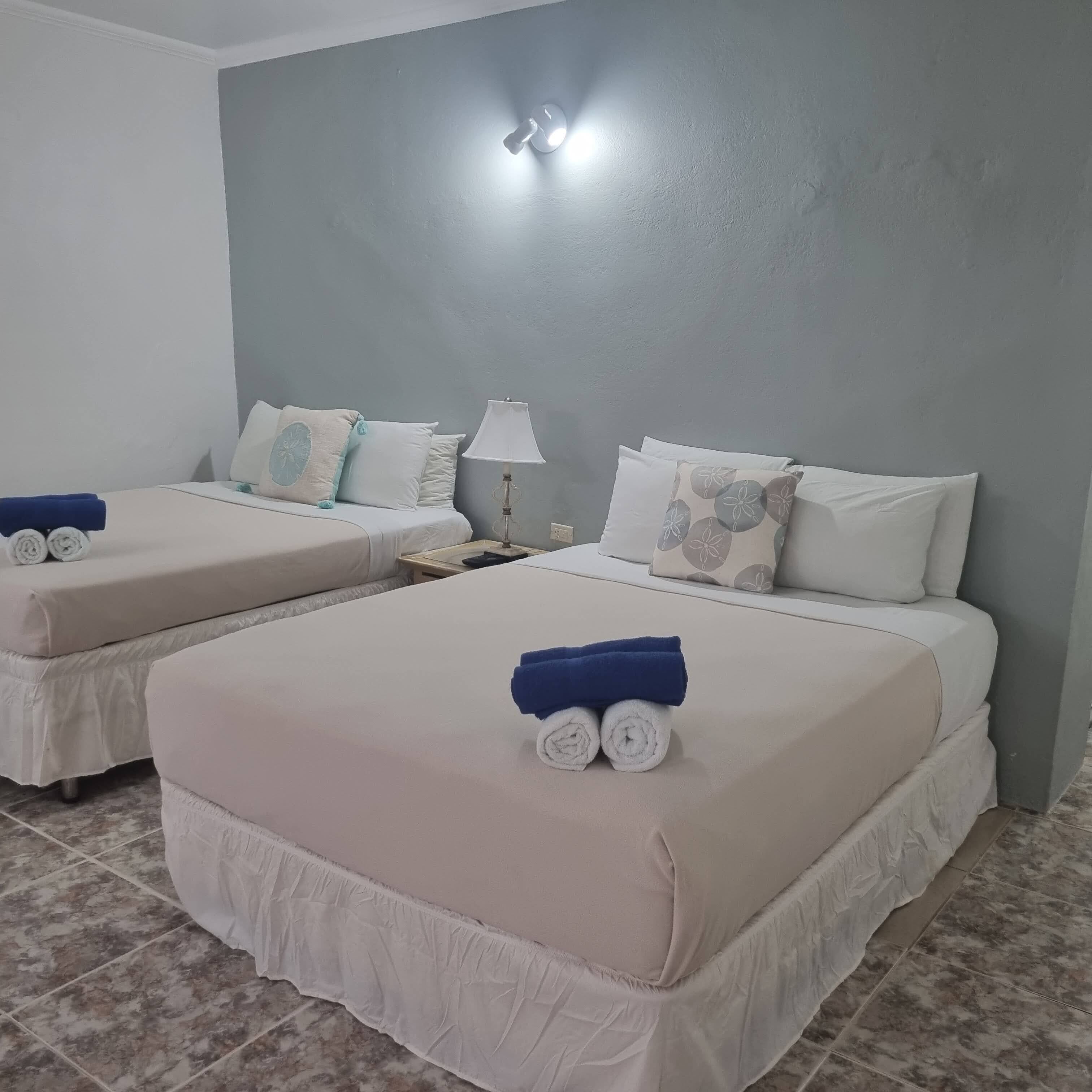 Een hotelkamer met twee bedden, elk met een witte sprei en blauwe handdoeken. Er staat een nachtkastje met een lamp tussen de bedden.