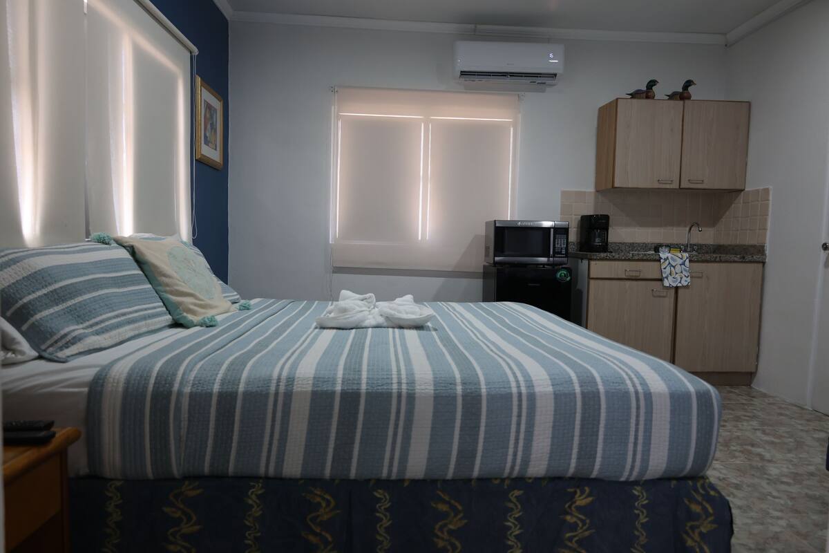 A bedroom with a bed, a bedside table, a microwave, a coffee maker, and a small kitchenette.
