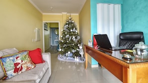 A living room with a Christmas tree, a sofa with a red pillow, a desk with a laptop, and a chair.