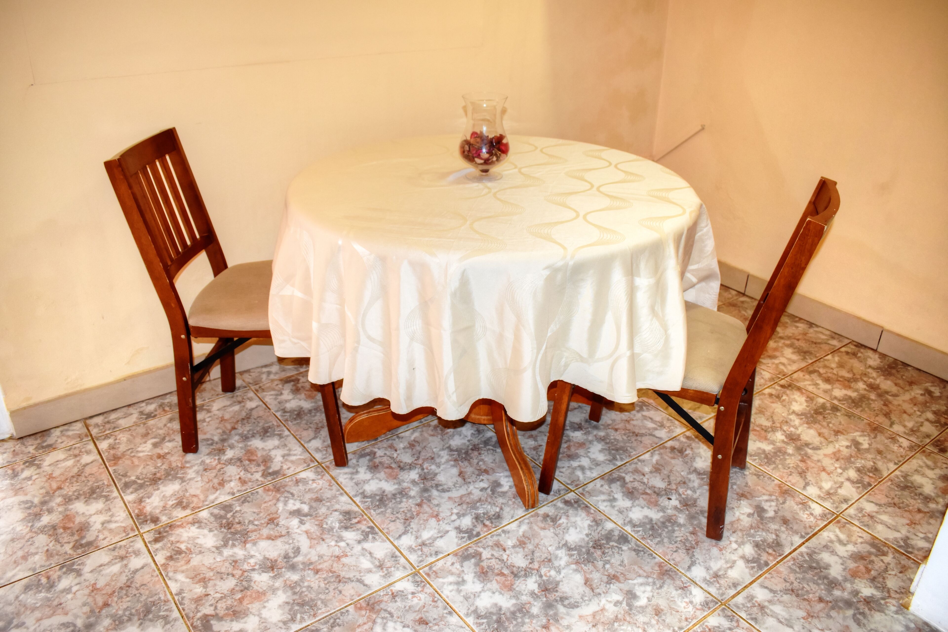 A round table with a white tablecloth, two wooden chairs, and a vase with flowers on the table.