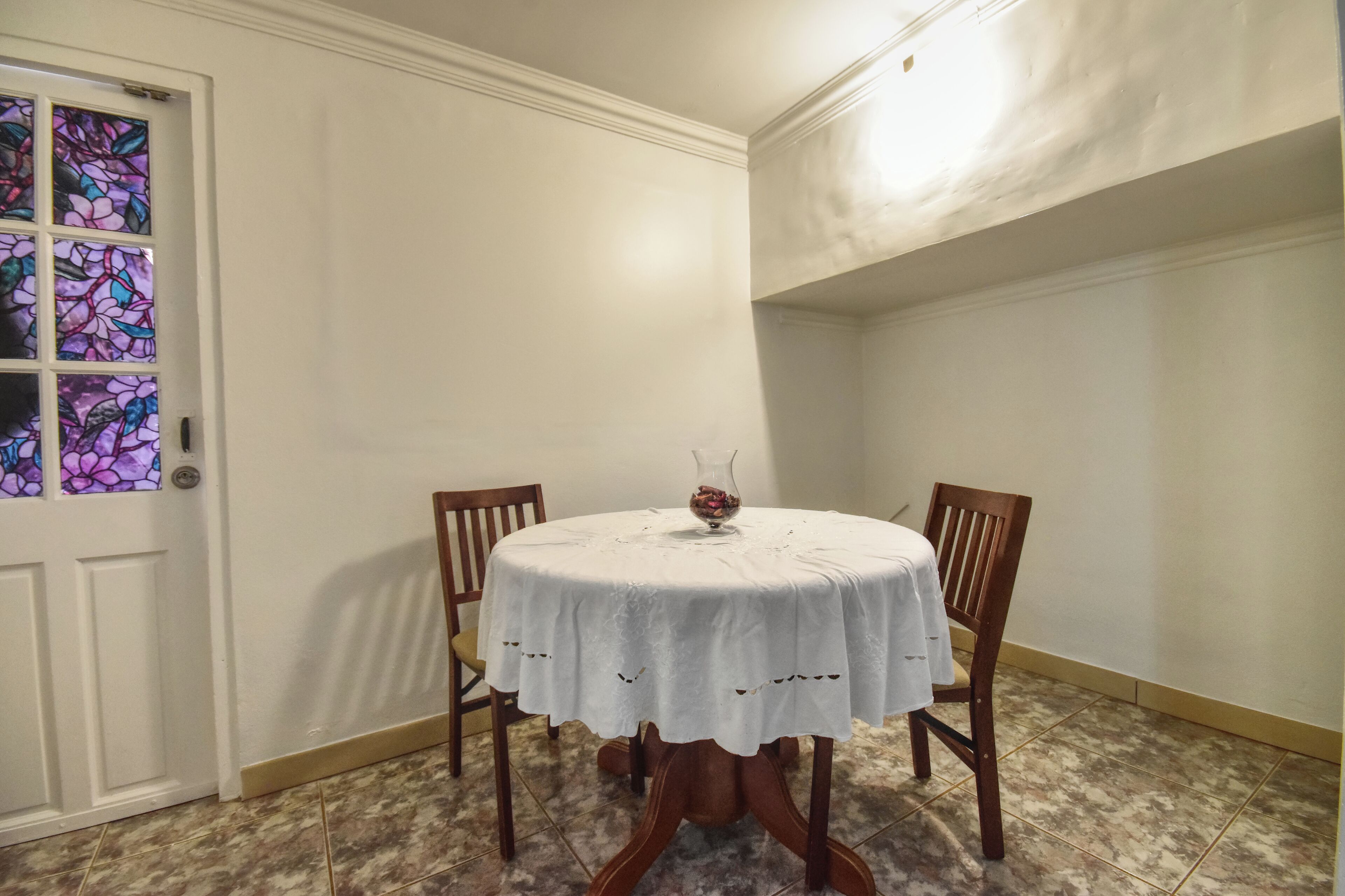 A dining area with a round table covered in a white tablecloth, two wooden chairs, and a vase with flowers on the table.