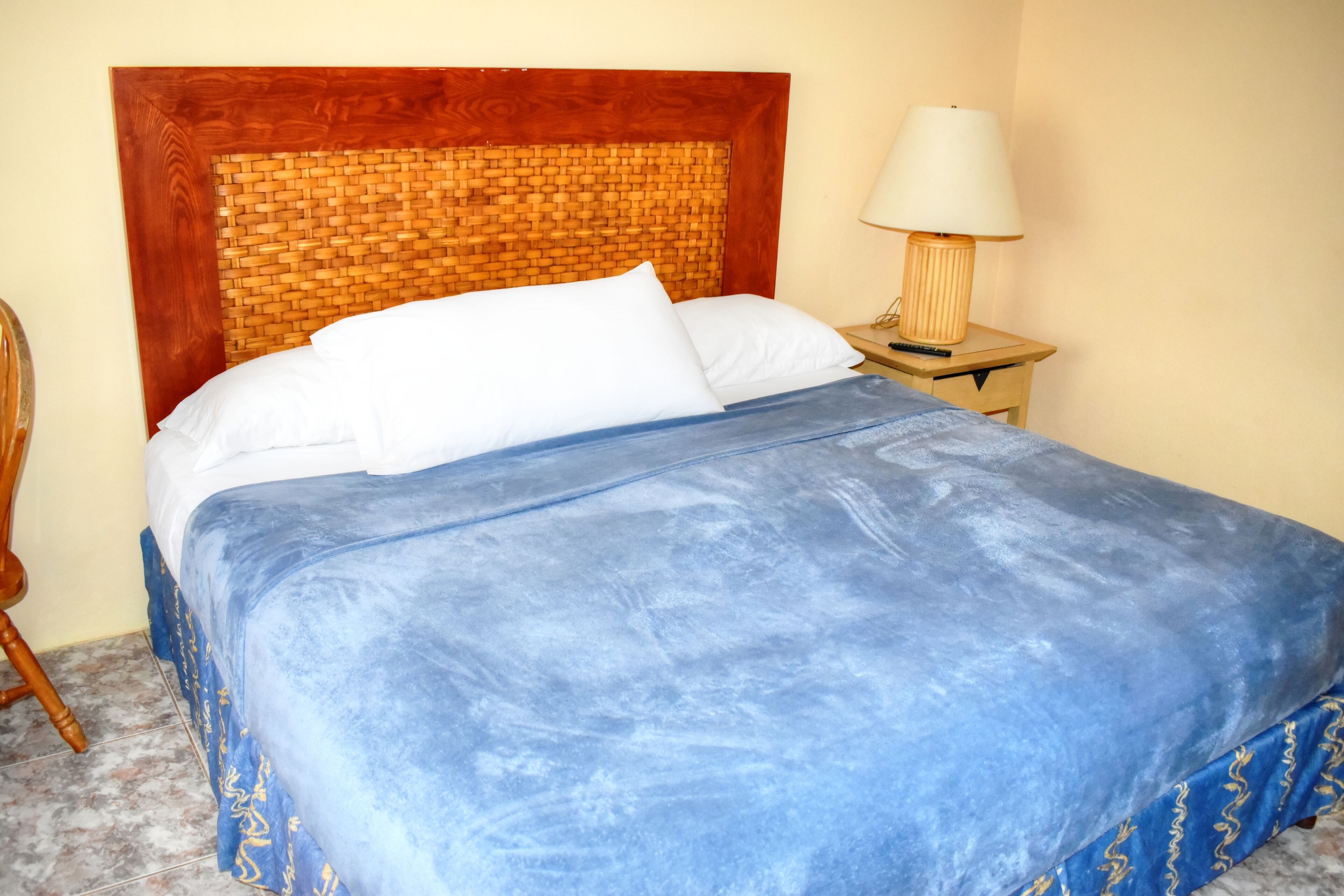 A bed with a blue bedspread and white pillows, a wooden headboard, and a nightstand with a lamp.