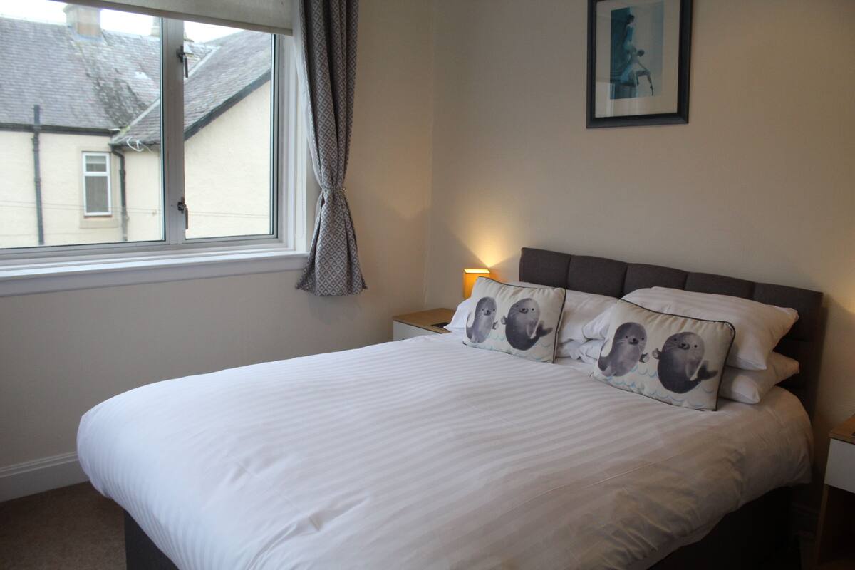 A neatly made bed with white linens and two pillows featuring sea animals. A bedside lamp is on the nightstand beside the bed.