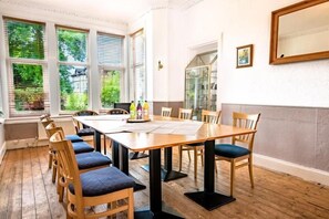 A dining area with a wooden table and chairs, large windows with blinds, and a view of greenery outside. - The Woodhouse Hotel (Largs)