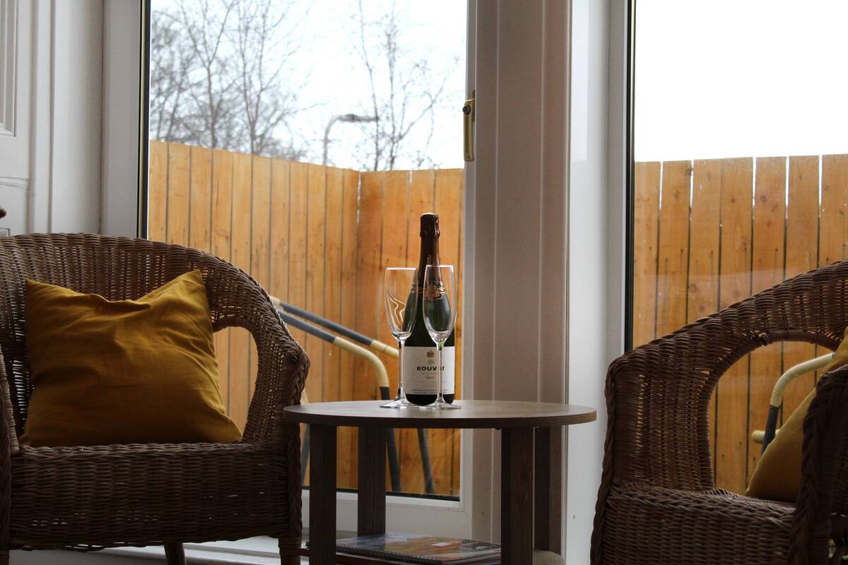 A wicker chair with a mustard-colored pillow, a small table with a bottle of wine and two glasses, and a wooden fence in the background.
