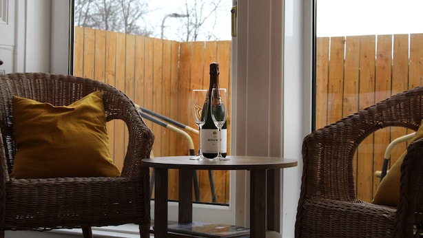 A wicker chair with a mustard-colored pillow, a small table with a bottle of wine and two glasses, and a wooden fence in the background. - The Woodhouse Hotel (Largs)