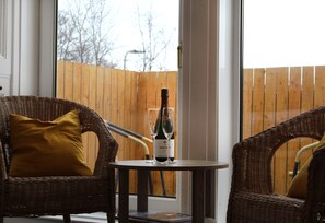 A wicker chair with a mustard-colored pillow, a small table with a bottle of wine and two glasses, and a wooden fence in the background. - The Woodhouse Hotel (Largs)