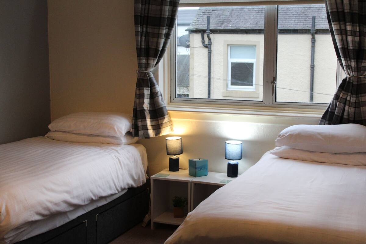 A bedroom with two single beds, a nightstand with two lamps, and a window with curtains.