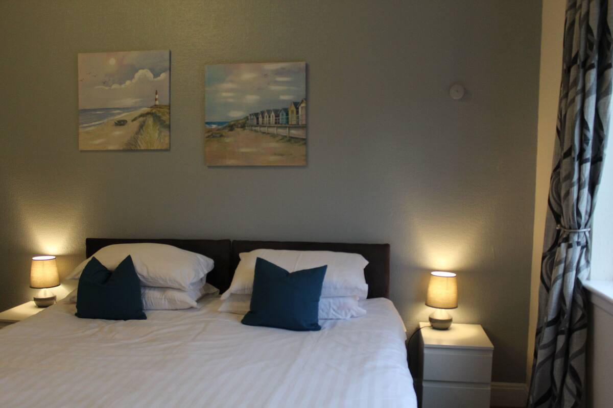 A neatly made bed with white linens and two dark blue pillows, flanked by two bedside lamps on white nightstands.