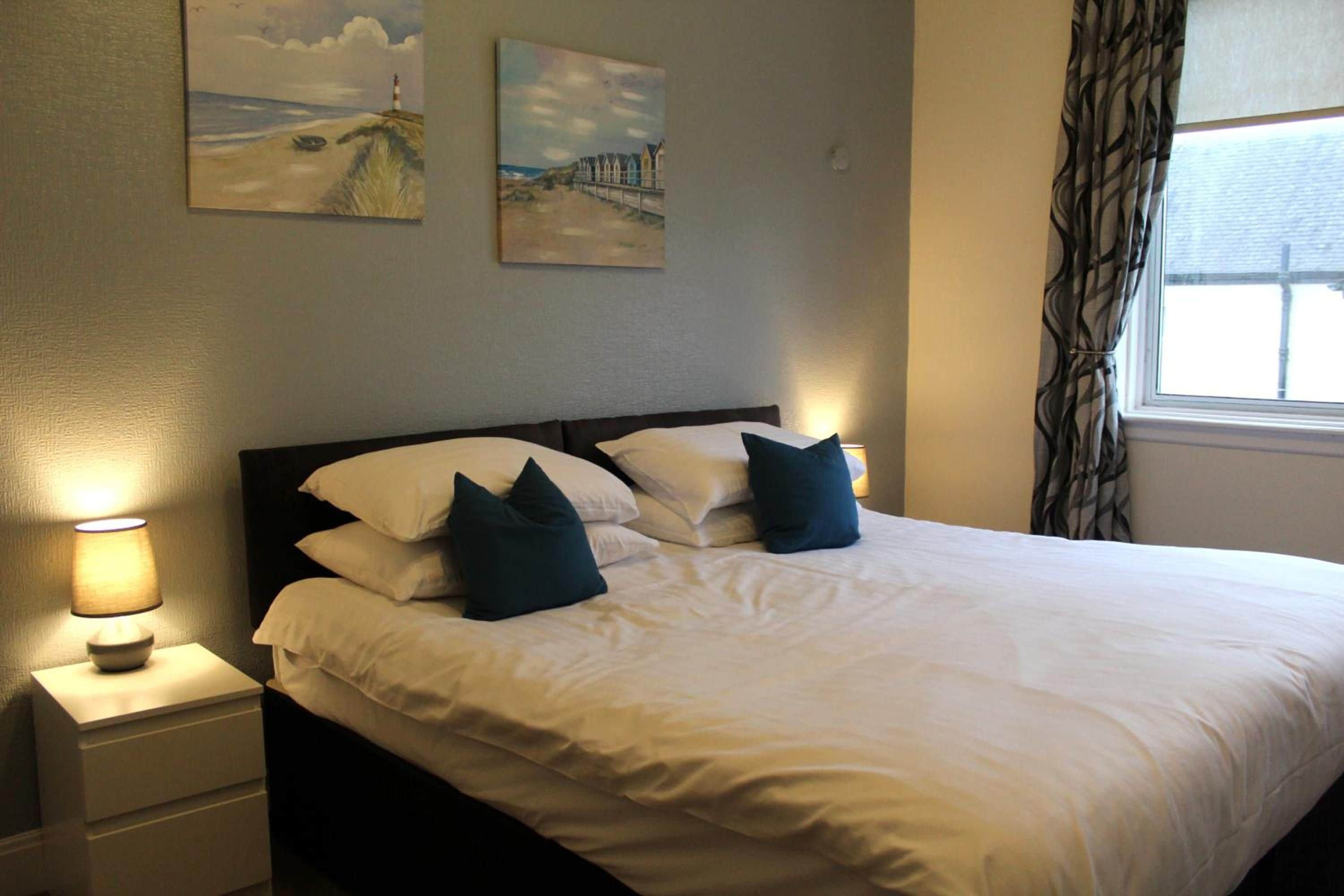 A neatly made bed with white linens and dark pillows, a bedside lamp, and two wall-mounted paintings depicting coastal scenes.