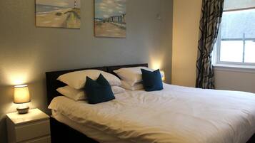 A neatly made bed with white linens and dark pillows, a bedside lamp, and two wall-mounted paintings depicting coastal scenes.