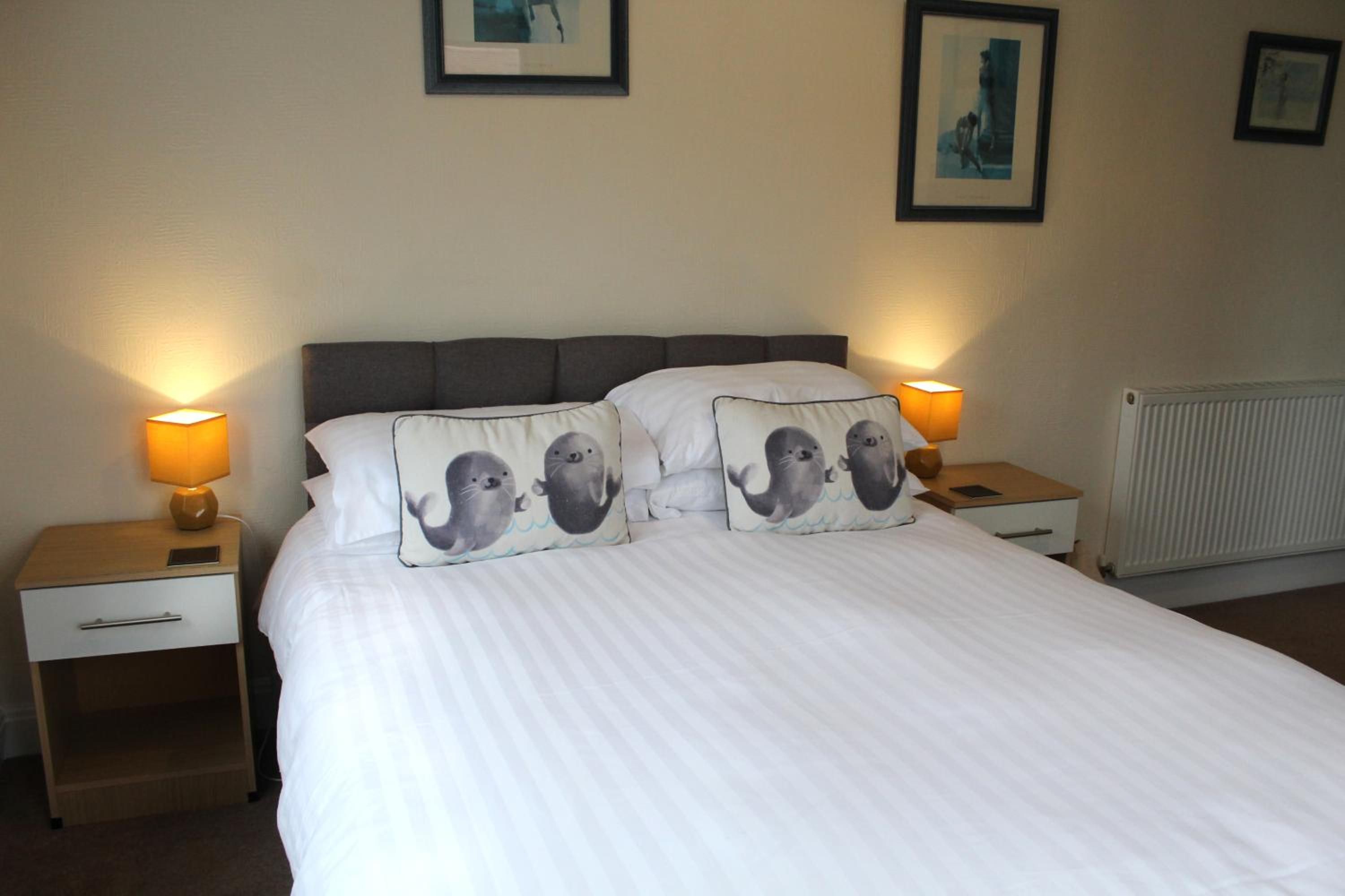 Una cama bien hecha con ropa blanca y dos almohadas con motivos de animales, flanqueada por mesitas de noche con lámparas.