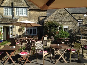 An outdoor seating area with tables and chairs, stone walls, and a large umbrella providing shade.