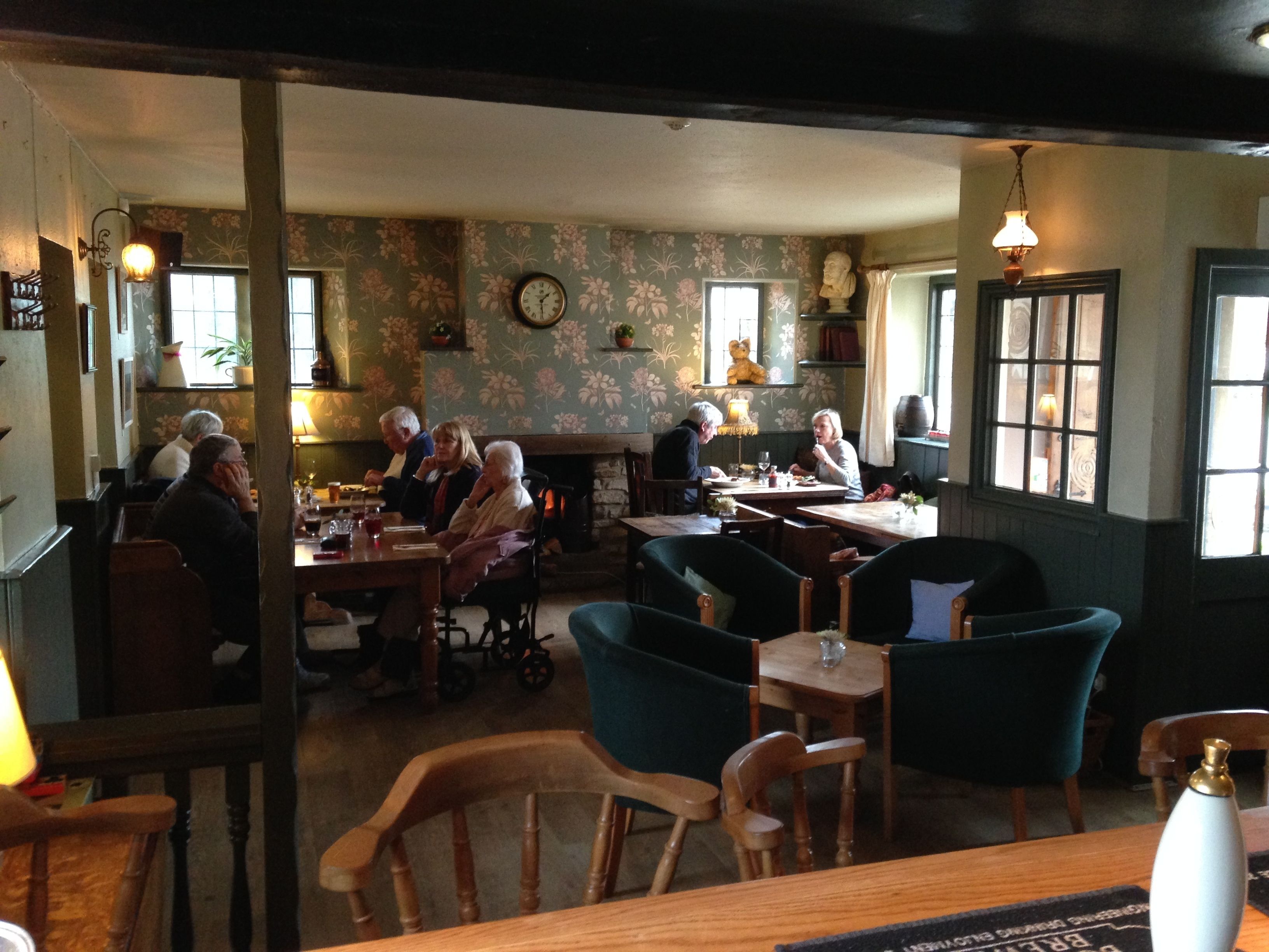 A cozy restaurant with wooden furniture and floral wallpaper. There are several tables with diners, and a clock is visible on the wall.