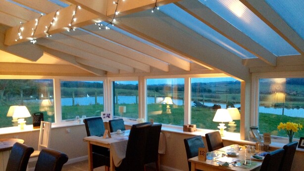 A dining area with a table set for two, chairs, and a view of a lake through large windows. - The Grainary Farm Stay (Scarborough)