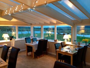 A dining area with a table set for two, chairs, and a view of a lake through large windows. - The Grainary Farm Stay (Scarborough)