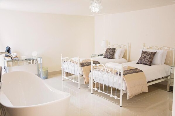 A hotel room with a white bathtub, two beds with white metal frames, a bedside table with a lamp, and a mirror.
