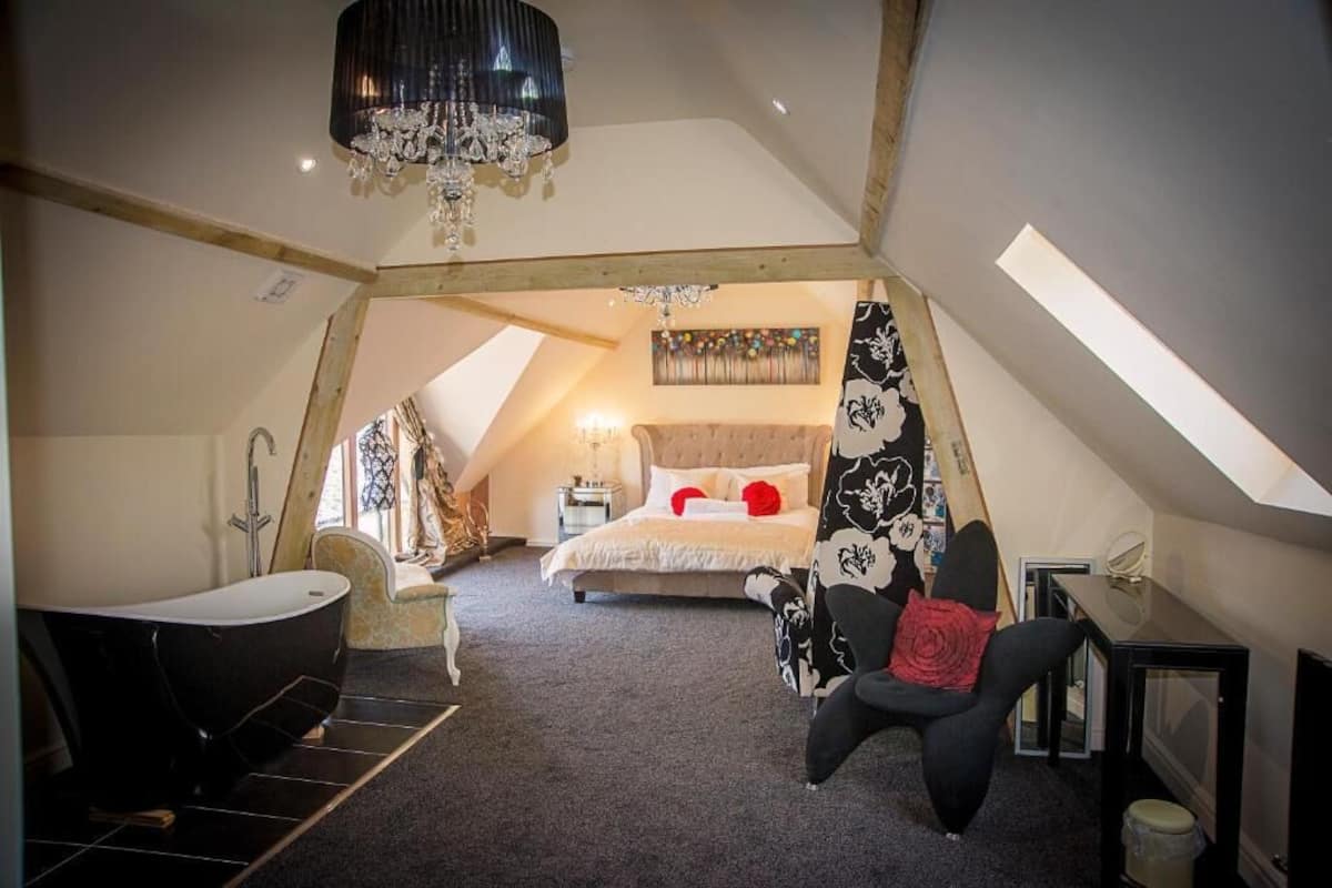 Un dormitorio con techo inclinado, una bañera negra, una cama con almohadas rojas, un sillón con estampado blanco y negro y una araña.