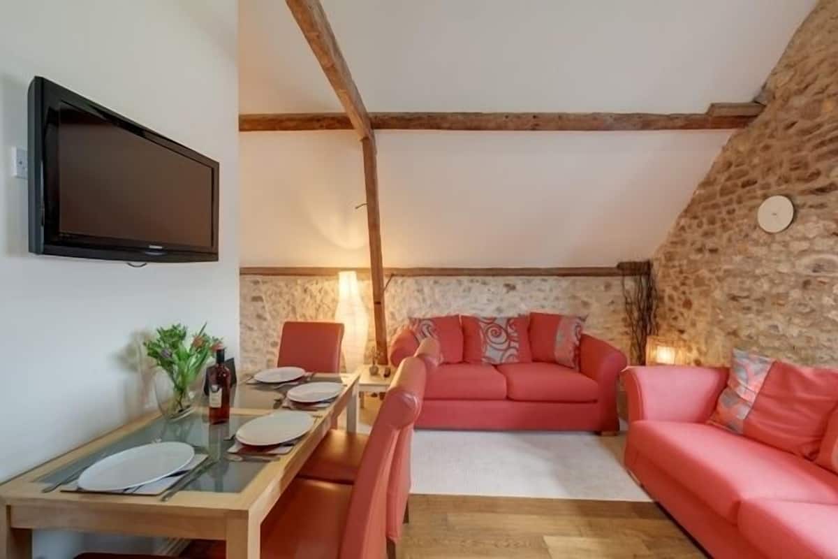 Un salón acogedor con pared de piedra, un sofá rosa, una mesa de comedor con sillas y un televisor de pantalla plana montado en la pared.