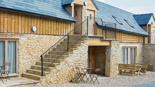 A stone building with wooden exterior walls, a staircase leading to a balcony, and outdoor seating. - HILLS FARM STAYS (Axminster)