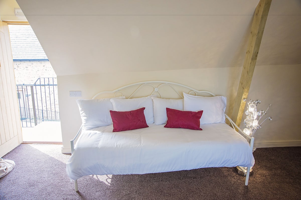 Una cama con ropa blanca y dos almohadas rojas, un trave de madera en el techo y vista al balcón exterior.