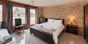 A bedroom with a stone wall, a bed, a chair, and a nightstand with a lamp.