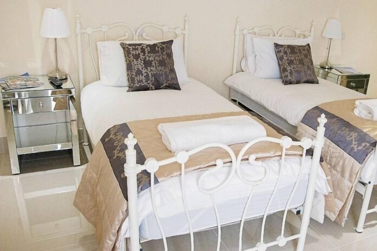 Habitación de hotel con dos camas, cada una con estructura metálica blanca y cubrecama color beige. Hay dos mesitas de noche con lámparas, y una mesita auxiliar con una lámpara y una revista.