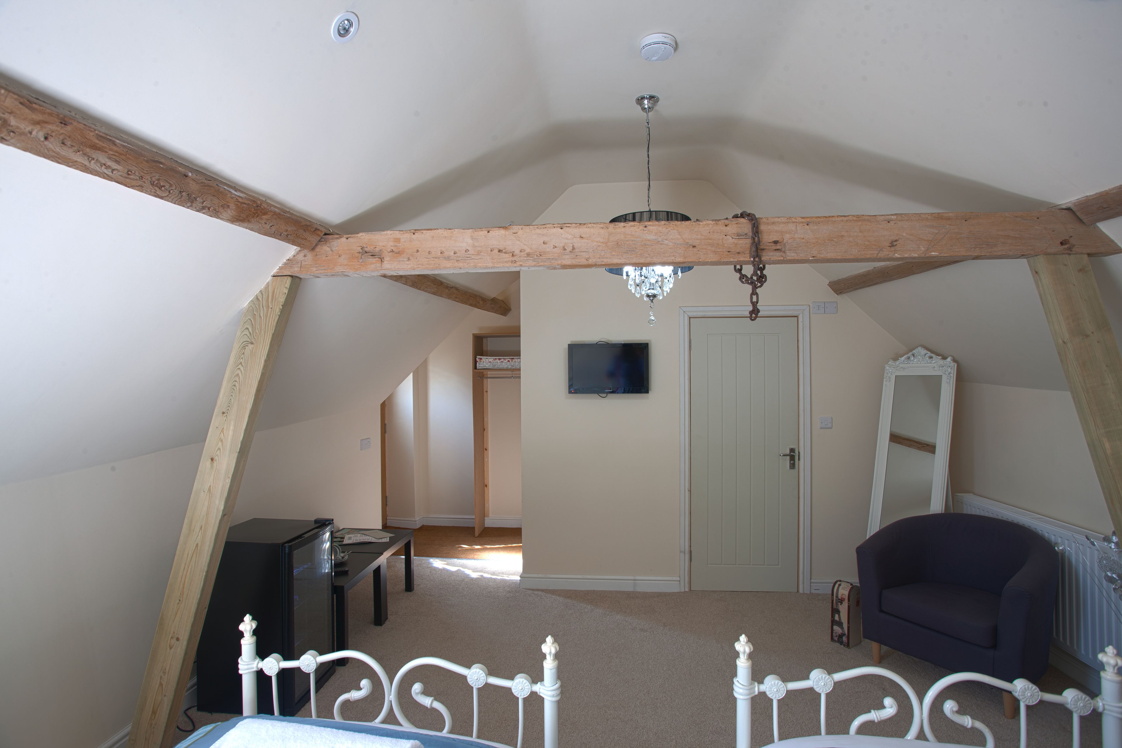 Un dormitorio en el ático con vigas de madera a la vista, un candelabro, una cama, una silla, un televisor y un espejo.